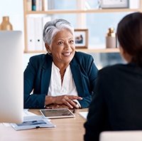 Woman with gray hair in blue blazer smiling at co-worker over desk